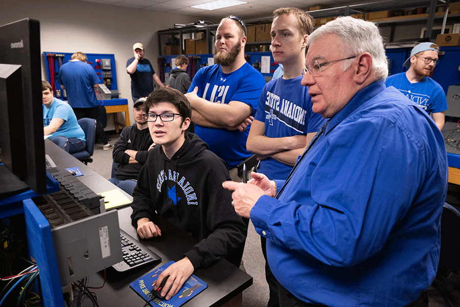 A white, older professor with white hair and glasses, and wearing a blue windbreaker jacket, stands next to two white male students in a control room as a third white male student sits at a computer terminal. The two standing students have short, blondish-brown hair and glasses and are wearing blue Indiana State t-shirts. The one further away from the instructor also wears a beard. The seated student has dark hair and glasses and wears a black Indiana State hoodie with blue letters on it.