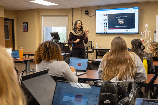 Female professor with dark hair and glasses standing in front of a classroom with back to camera. In the background is a human skeleton on a stand and a video board with medical information.