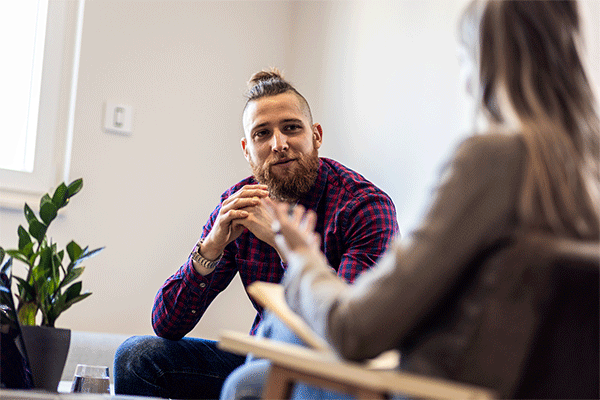 A white male with a beard and hair in a bun and facing the camera listens as a counselor in the foreground with her back to the camera and slightly out of focus speaks. 